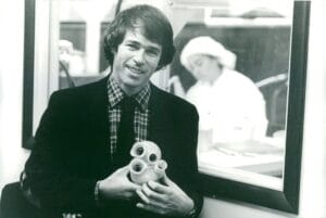 A young man in a suit and plaid shirt smiles, holding a mechanical heart device. Behind him, someone in a lab coat works in a lab, seen through a window.