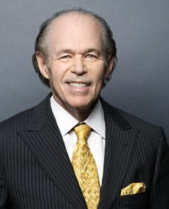 An older man with thinning gray hair, in a dark pinstripe suit, white shirt, and gold patterned tie, poses with a smile against a plain gray background.