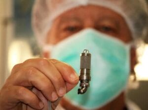 A person in a medical mask and hair net holds a small metallic instrument up close, their blurred face in the background.