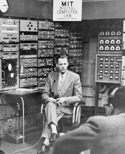 A man in a suit sits in front of a large panel of early computer equipment at the MIT Digital Computer Lab, with wires, switches, and dials visible on the machines behind him.