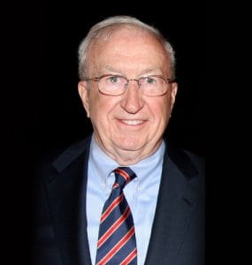 An older man with glasses, gray hair, and a friendly smile in a dark suit and striped tie stands before a plain black background.
