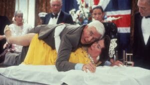 A man in a suit falls onto a woman in a yellow dress and tiara on a table, with shocked guests watching at a formal event.