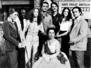 Seven people stand behind a seated woman dressed as a queen with crown and sash; brick wall and SAVE GREAT BRITAIN sign in back. Comedy skit.