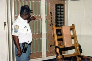 A corrections officer shows us a prison's empty wooden electric chair. He wears a uniform, cap, and radio.