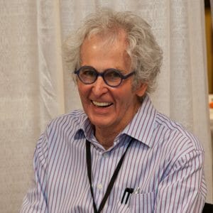 An older man with curly gray hair, blue-rimmed glasses, and a striped shirt smiles seated before white curtains, black lanyard around neck.