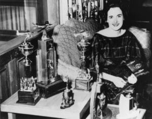 A smiling woman in a plaid dress sits on a patterned couch, trophies around her on the table and floor, vintage black-and-white scene.