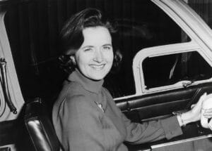 A woman with dark hair smiles from the pilot seat of a small plane. She wears a long-sleeved top, bracelet, and the window vent is open.