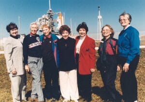 Seven women smiling together in front of a space shuttle launch pad on a sunny day, with the shuttle and blue sky behind them.