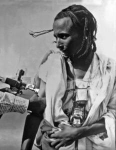 A man with braided hair and traditional clothing receiving an injection in their upper arm from someone holding a jet injector. The image is black and white.