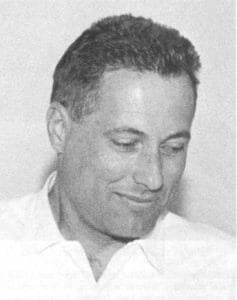 Black and white portrait of a man with short, dark hair in a light collared shirt, gazing downward with a faint smile.
