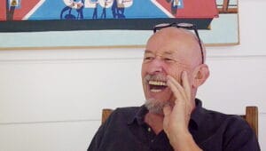 A bald man with a beard and glasses on his head laughs while seated by a white wall with a colorful painting in the background.