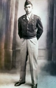 Black and white photo of a young man in uniform, hands behind back, in front of a studio backdrop; jacket, tie, and cap visible.