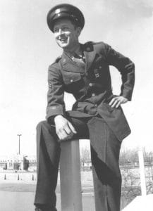 A young man in a World War II military uniform and cap sits on a post outdoors, smiling and looking into the distance. Trees and buildings can be seen in the background under a clear sky.
