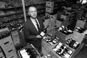 A man in a suit stands holding a wine bottle in a cluttered grocery store with stacked bottles, boxes, and product-filled shelves behind him.