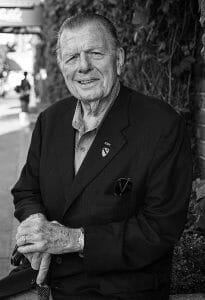 An elderly man in a dark suit jacket stands outdoors, smiling gently with a cane. Ivy-covered walls behind him. Black and white photo.