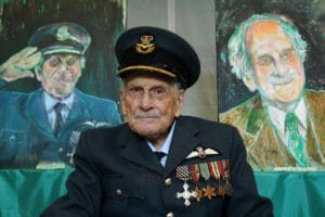 An elderly veteran in uniform, with medals and a cap, sits before two portraits: one in military attire, one as an older civilian.