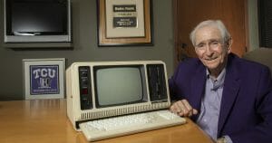 An elderly man in a purple jacket smiles at a desk with a TRS-80 computer; plaques and a TCU Honors College sign are behind him.