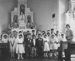 A group of solemn children in formal attire, some in white dresses and veils, pose by a church altar with statues; a stern-looking adult woman stands nearby.