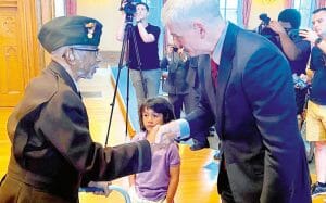 An elderly man in a military uniform shakes hands with a suited man as a child stands between them; people photograph the scene.