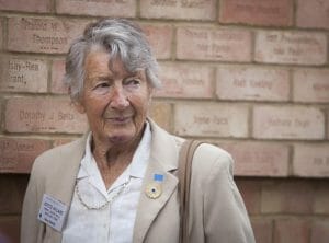An older woman with gray hair in a beige blazer stands before a wall engraved with names, smiling slightly, wearing a gold necklace.