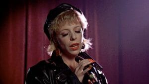 A woman with short blonde hair, black leather jacket, and cap sings into a mic on stage before red curtains, pink-purple lights.