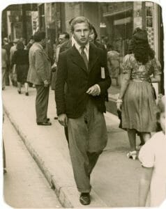 A young man in a suit jacket and tie walks down a busy city sidewalk holding a book; others are in the background. Black and white vintage photo.