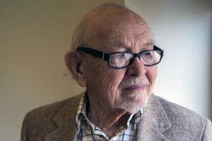 An elderly man with glasses and a gray beard looks to the side, in a plaid shirt and brown jacket by a softly lit wall.
