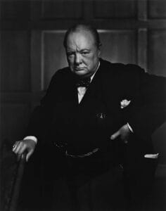 A stern-looking Winston Churchill in a dark suit and bow tie stands with one hand on a chair and the other on his hip, posed against a wooden paneled wall in a classic black-and-white portrait.