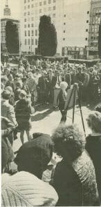 A large crowd gathers outdoors as a woman stands painting at an easel in a city plaza; 1950s buildings and BLUMS sign behind.