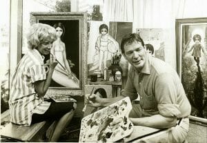 A man with a paint palette paints in a studio of big-eyed portraits as a woman sits nearby, smiling and watching him.
