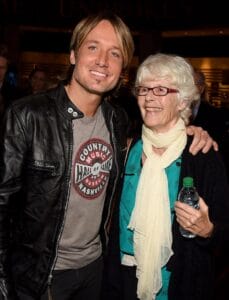 A smiling man in a black leather jacket stands by an older woman with white hair and glasses, holding a water bottle indoors.