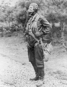 A Korean War-era pilot outdoors in full flight gear, including helmet and mask, with trees and a gravel path behind him.