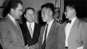 Four men in suits stand indoors. The man on the left, Vice President Nixon, shakes hands with the center man, who is Korean; two others, one in glasses and a bow tie, watch.