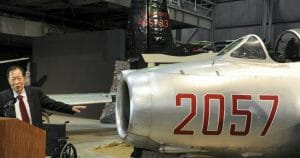 An older man in a suit speaks at a lectern beside a silver Russian jet fighter numbered 2057, inside an aviation museum hangar.