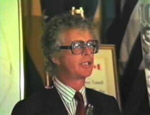 A person with curly gray hair and large glasses is speaking at an event, dressed in a striped shirt, tie, and suit. There are flags and a framed picture in the background.