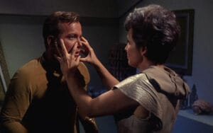 A woman reaches out and touches a mans face with both hands in a tense moment inside a futuristic room, with both characters wearing sci-fi style clothing.