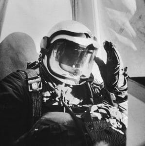 A person in a full helmet and pressurized suit sits in a high-altitude craft, one gloved hand raised, sunlight shining brightly.
