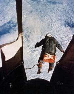 A helmeted person leaps from a balloon capsule high above Earth, clouds and terrain below, in free fall with a visible cord for a parachute trailing behind.