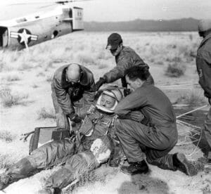 Four men in military uniforms help a man in a pressure suit sitting on desert ground near parachute lines, with a U.S. helicopter nearby.