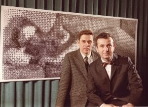 Two men in suits stand before a large artwork of a nude, created by digital means, set against a green curtain background.