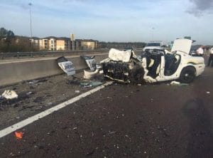 A heavily damaged sheriffs patrol car sits on a highway shoulder, its front end completely destroyed and debris scattered across the road. Other vehicles and people are visible in the background.