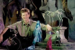 A man smiles among colorful skeleton and ghost puppets in a spooky animated graveyard with tombstones, eerie trees, and a skeletal horse.