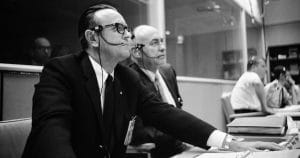 Two men in suits and headsets sit at consoles in a control room, focused on monitors and paperwork, likely during a mid-20th-century space mission. Other people work in the background behind glass dividers.