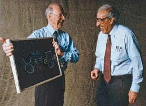 Two elderly men in dress shirts and ties laugh together; one holds a chalkboard with colorful drawings and the words hinge and base.
