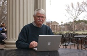 An older man with white hair and glasses sits outside at a table with a laptop, near large columns and a campus in the background.