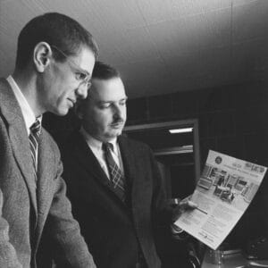Two men in suits stand indoors; one points while the other examines a GE-225 computer system brochure with images and text.