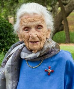 An elderly woman with white hair, blue top, floral scarf, and gold chain with a red medal stands outdoors among green trees.