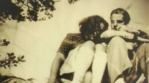 Sepia photo of two people sitting close outdoors, a woman leaning on the man's shoulder under tree branches; nostalgic vintage vibe.