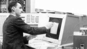 A man uses a light pen to interact with an early computer screen displaying graphics, sitting at a vintage computer terminal with control panels and buttons in the background.
