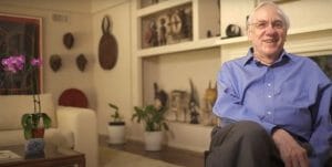An older man in a blue shirt smiles in a cozy living room with plants, African masks, artifacts on shelves, and a purple orchid nearby.